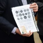 Человек держит лист с изображением монет, одет в темный костюм.