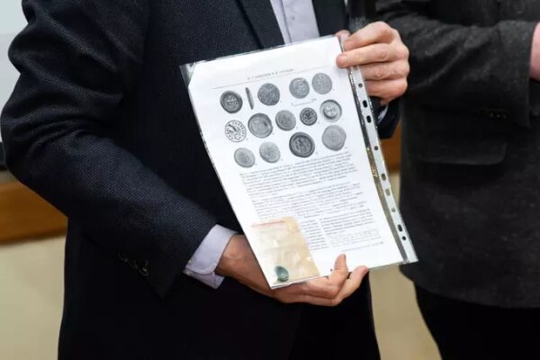 Человек держит лист с изображением монет, одет в темный костюм.