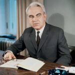 Мужчина в костюме сидит за столом с открытой книгой и очками в руке, светлая комната.