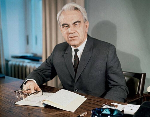 Мужчина в костюме сидит за столом с открытой книгой и очками в руке, светлая комната.