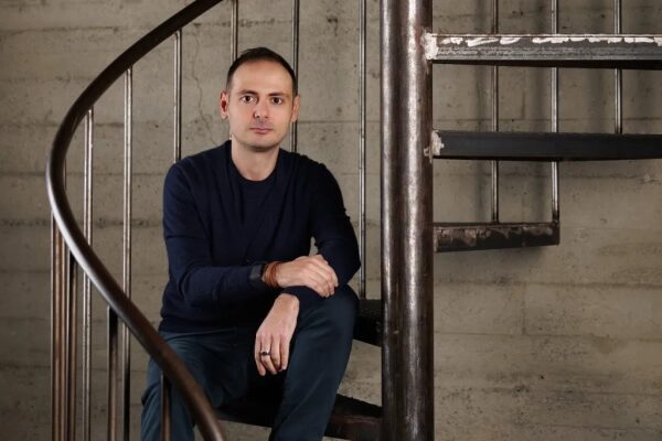 Мужчина сидит на металлической винтовой лестнице в помещении в темной одежде.