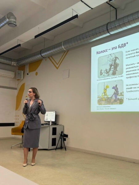 Презентация в офисе: женщина в костюме выступает перед экраном с слайдом о Колоссе.