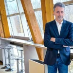 Мужчина в деловом костюме стоит в современном офисе с большими окнами.