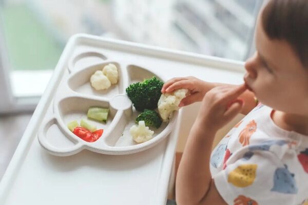 Ребенок ест овощи из тарелки с разделителями, включая брокколи и цветную капусту.