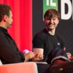 Два человека обсуждают на технологической конференции, сидя на сцене.