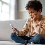 Ребёнок сидит на диване с телефоном, задумчиво глядя на экран.