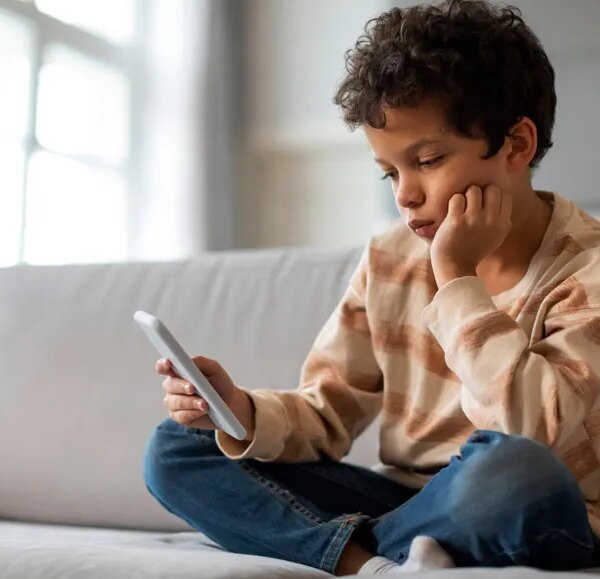 Ребёнок сидит на диване с телефоном, задумчиво глядя на экран.