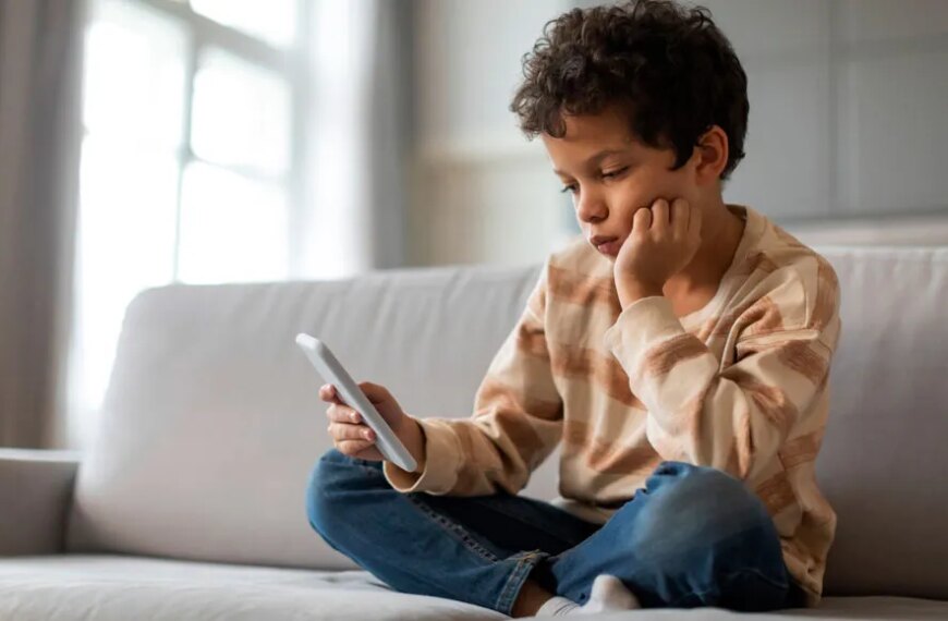 Ребёнок сидит на диване с телефоном, задумчиво глядя на экран.