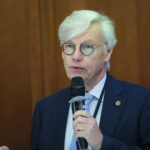 Мужчина в очках и костюме говорит в микрофон на фоне деревянной стены.