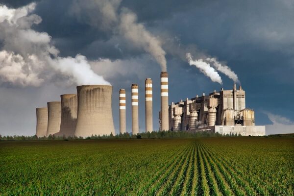 Фабрика с трубами, выбрасывающими дым, на фоне поля и неба.