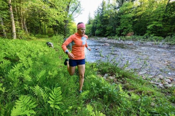 Женщина в спортивной одежде бежит по лесной тропе рядом с речкой, весенний пейзаж.