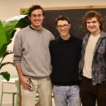 Трое мужчин позируют у доски с формулами, растением на фоне.