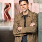 Мужчина в коричневой куртке стоит, скрестив руки, у стеклянной стены внутри здания.