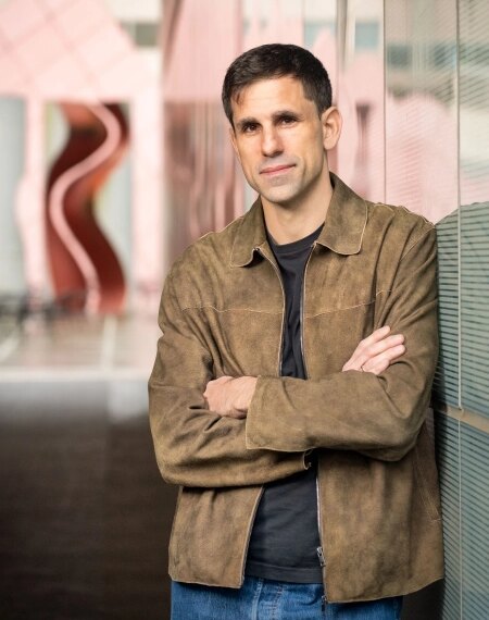 Мужчина в коричневой куртке стоит, скрестив руки, у стеклянной стены внутри здания.