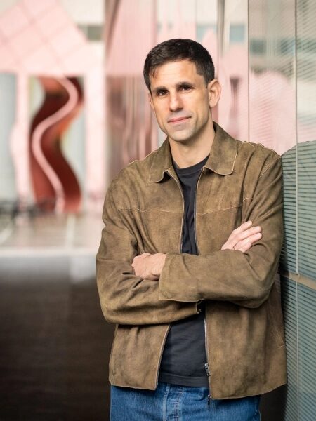 Мужчина в коричневой куртке стоит, скрестив руки, у стеклянной стены внутри здания.