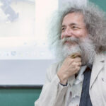 Мужчина с седой бородой и кудрявыми волосами улыбается на фоне презентации.