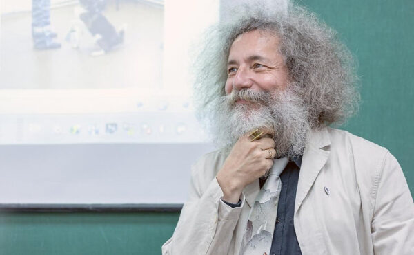 Мужчина с седой бородой и кудрявыми волосами улыбается на фоне презентации.