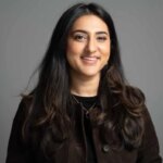 Женщина с длинными волосами в темной одежде, улыбается на сером фоне.