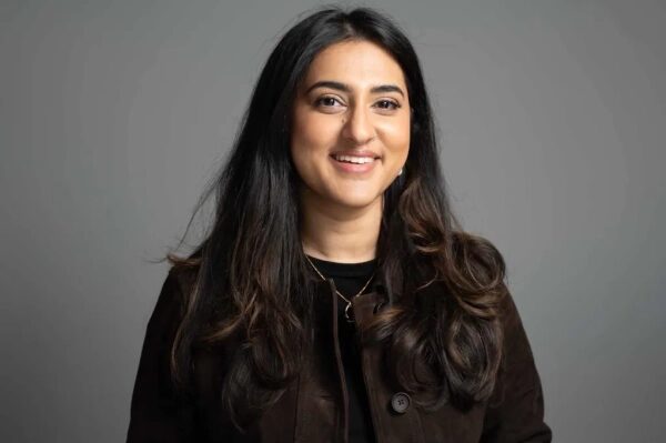 Женщина с длинными волосами в темной одежде, улыбается на сером фоне.