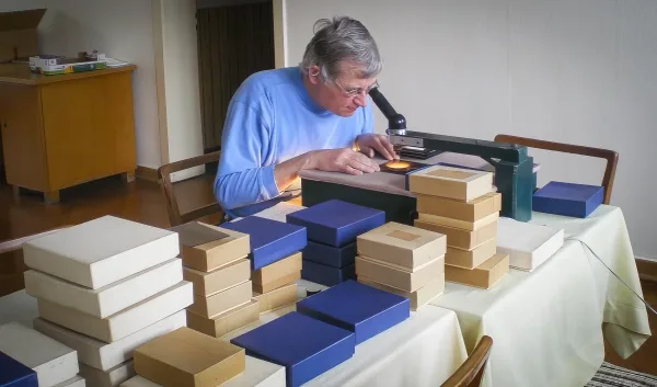 Мужчина, окруженный коробками с униформой, рассматривает фотографию на стеклянной пластине с помощью светового планшета и увеличительного стекла.