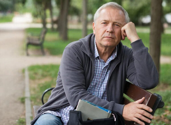 Человек в парке сидит на скамейке с книгой на коленях, расслабленная обстановка.