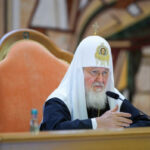 Патриарх в белом клобуке говорит за столом, микрофон, церковь.