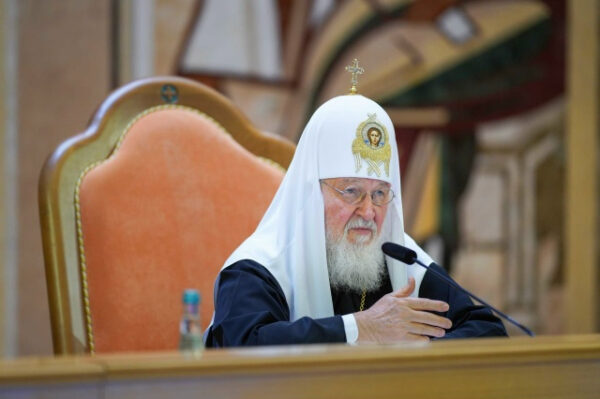 Патриарх в белом клобуке говорит за столом, микрофон, церковь.