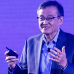 Мужчина в костюме выступает с микрофоном на фоне фиолетового экрана.