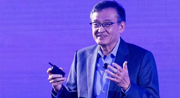 Мужчина в костюме выступает с микрофоном на фоне фиолетового экрана.