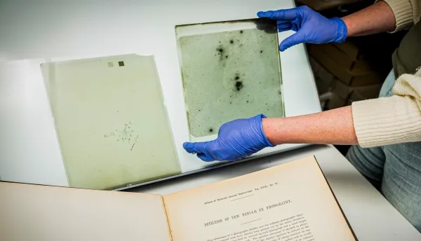 В перчатках руки держат квадратную стеклянную фотопластинку с пометками, сделанными маркером, а другая рука лежит на световом коробе позади.