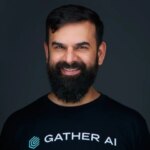 Мужчина с бородой в футболке Gather AI на темном фоне, улыбается.