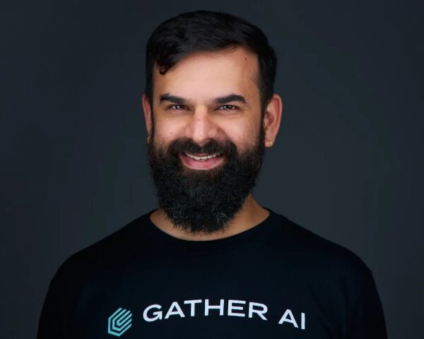 Мужчина с бородой в футболке Gather AI на темном фоне, улыбается.