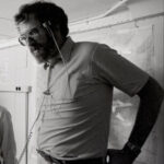 Человек в рубашке с гарнитурой разговаривает, стоя перед картой на стене.