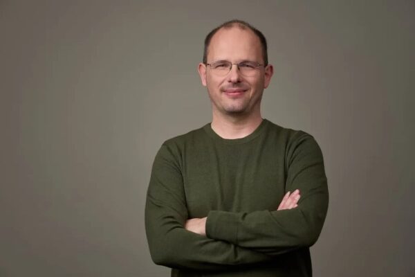 Мужчина в темно-зеленом свитере стоит, скрестив руки, на сером фоне.