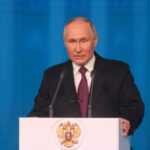 Мужчина в костюме выступает на трибуне с гербом России, фон синие полосы.