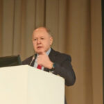 Человек выступает на конференции за трибуной, делая жест рукой.