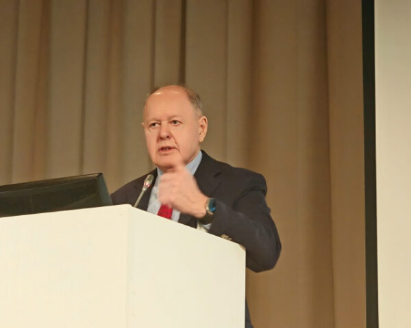 Человек выступает на конференции за трибуной, делая жест рукой.