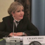 Женщина в деловой одежде за столом на конференции, микрофон и табличка "Сидоренко".