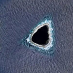 Треугольный остров в океане, окружённый темной водой и светлым берегом.