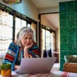 Пожилая женщина за ноутбуком дома в вязании, уютная обстановка.