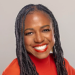 Женщина с дредами в красной блузке, улыбается на светлом фоне.