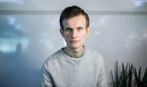 Мужчина в сером свитере на фоне белой стены и зелёного растения в офисе.