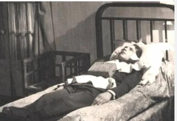 Человек в кровати, одет в костюм, с книгой на груди, комната в ретро-стиле.