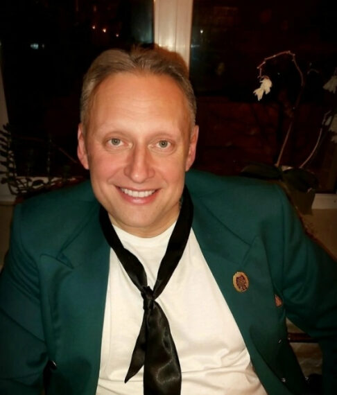 Мужчина в зеленом пиджаке с чёрным галстуком и значком на фоне окна.