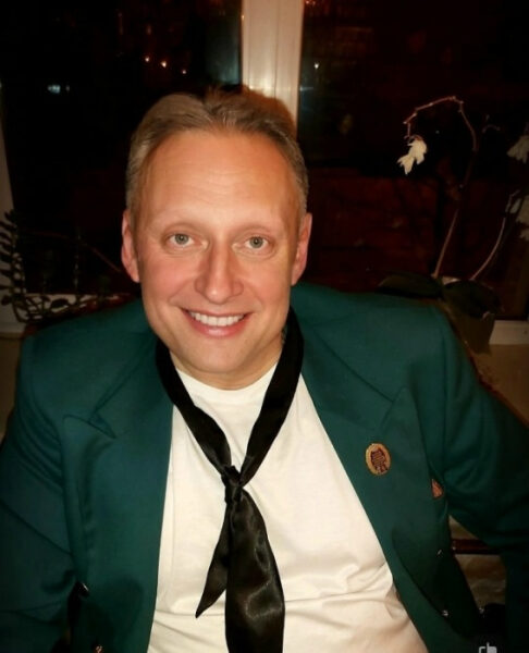 Мужчина в зеленом пиджаке с чёрным галстуком и значком на фоне окна.