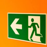 Указатель аварийного выхода на оранжевой стене, стрелка указывает налево.