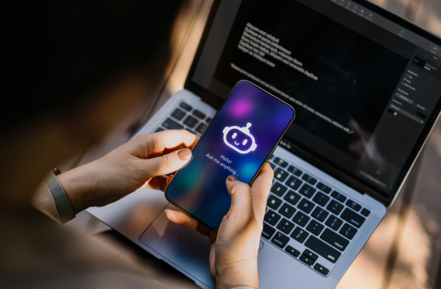 Человек держит смартфон с изображением чат-бота, на фоне открыт ноутбук с кодом.