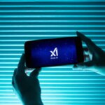 Смартфон с логотипом Grok AI в руках на фоне голубых жалюзи.