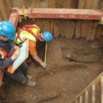 Археологи в касках исследуют раскопки с останками человека, делают записи.