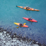 Три каяка с людьми на бирюзовой воде возле каменного берега.